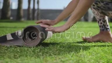 女子手在公园训练后卷起瑜伽垫。 女孩在草地上折叠垫子